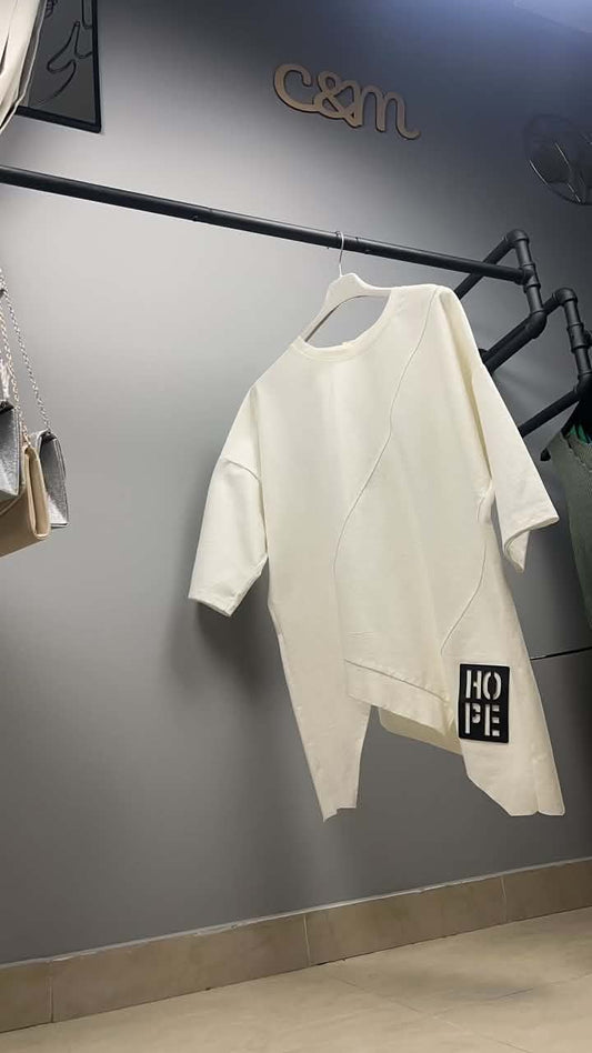 Η λευκή ασύμμετρη μπλούζα "HOPE" κρεμασμένη σε κατάστημα ρούχων. Φαίνεται η ιδιαίτερη γραμμή της, το φαρδύ μανίκι και η διαγώνια ραφή στο μπροστινό μέρος.